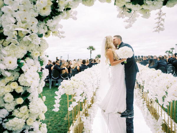 A bride and groom share a kiss at the end of a flower-lined aisle under a floral arch, surrounded by seated guests at an outdoor wedding ceremony.