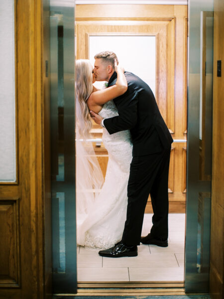 A bride and groom share a kiss in an elevator, framed by wooden doors. The bride wears a white, strapless wedding gown and veil, while the groom is in a black suit.