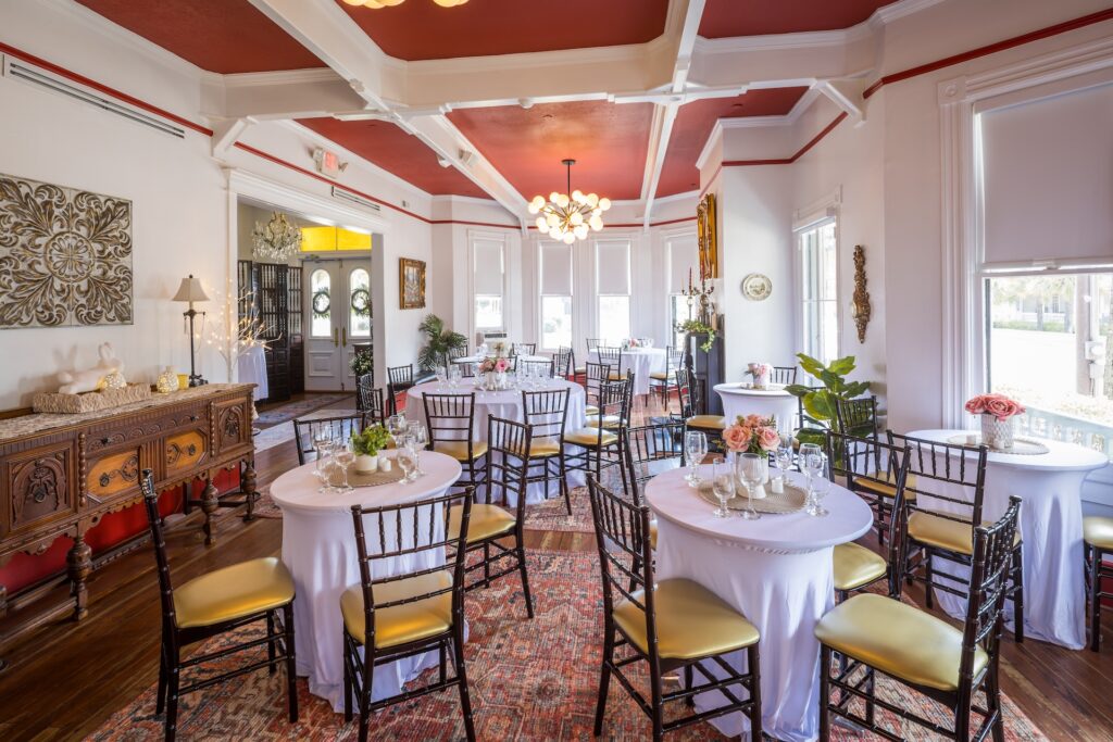 A bright, elegant dining room with round tables covered in white cloths, gold chairs, floral centerpieces, ornate sideboard, and large windows letting in natural light. The ceiling is red with white beams and chandeliers.