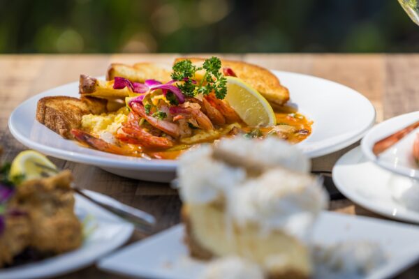 A plate of shrimp and grits garnished with parsley, red onion, and a lemon wedge, served with toasted bread on a wooden table; a blurred dessert appears in the foreground.