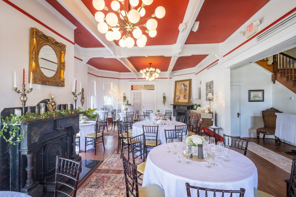 Elegant dining room with round tables set for an event, ornate chandeliers, red and white ceiling, vintage mirror above a fireplace, classic decor, wooden floors, and artwork on the walls. Natural light fills the space.