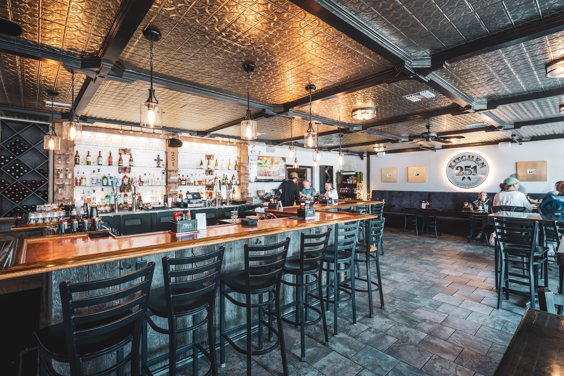 A modern bar and restaurant interior with a long wooden bar, high black chairs, bottles on shelves, and pendant lights. Several people are seated at tables in the background. The ceiling features a decorative metal pattern.