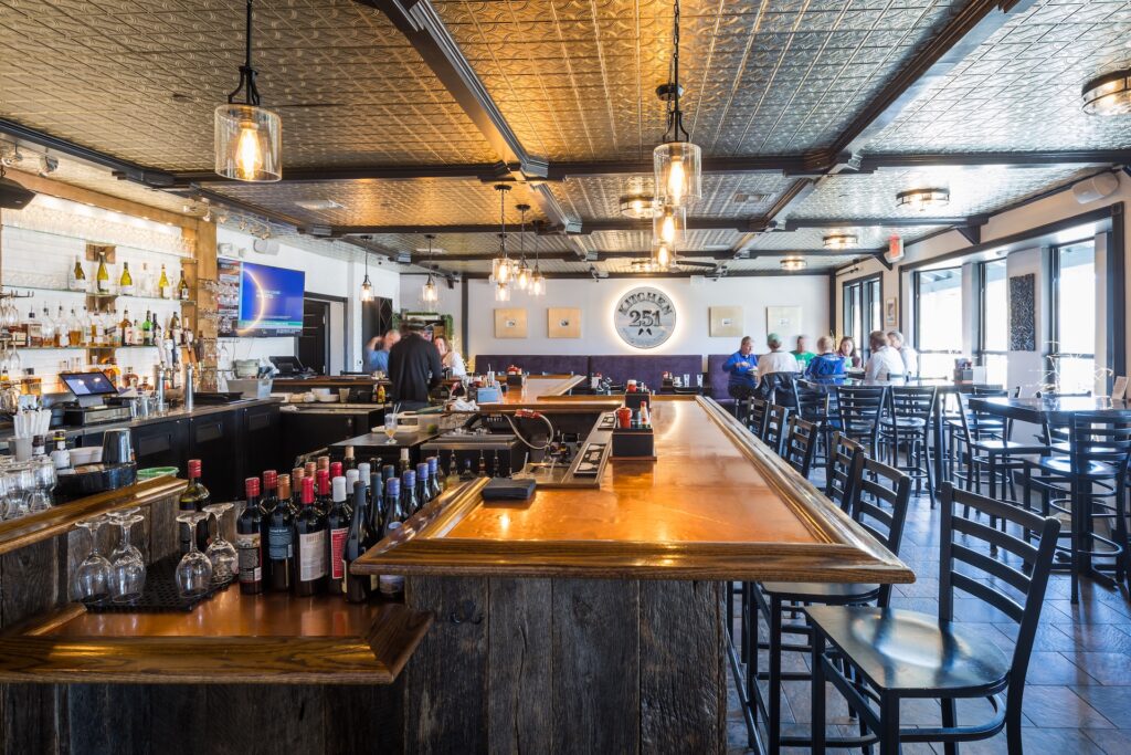 Spacious bar and restaurant with a wooden bar counter, bottles of wine and liquor, high tables with chairs, decorative ceiling, hanging lights, and large windows letting in natural light. A few people are seated near the windows.