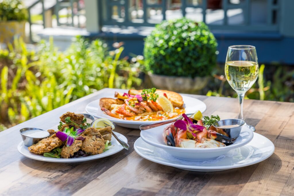 Three plates of gourmet food, including seafood and fried items, are served on a wooden outdoor table with a glass of white wine beside them. The table is set in a garden with greenery in the background.