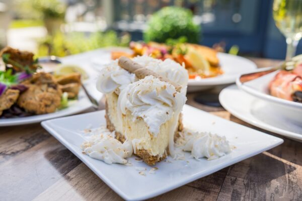 A slice of creamy cheesecake topped with whipped cream and a wafer stick is served on a white plate, garnished with toasted coconut. Other dishes and drinks are blurred in the background on a wooden table.