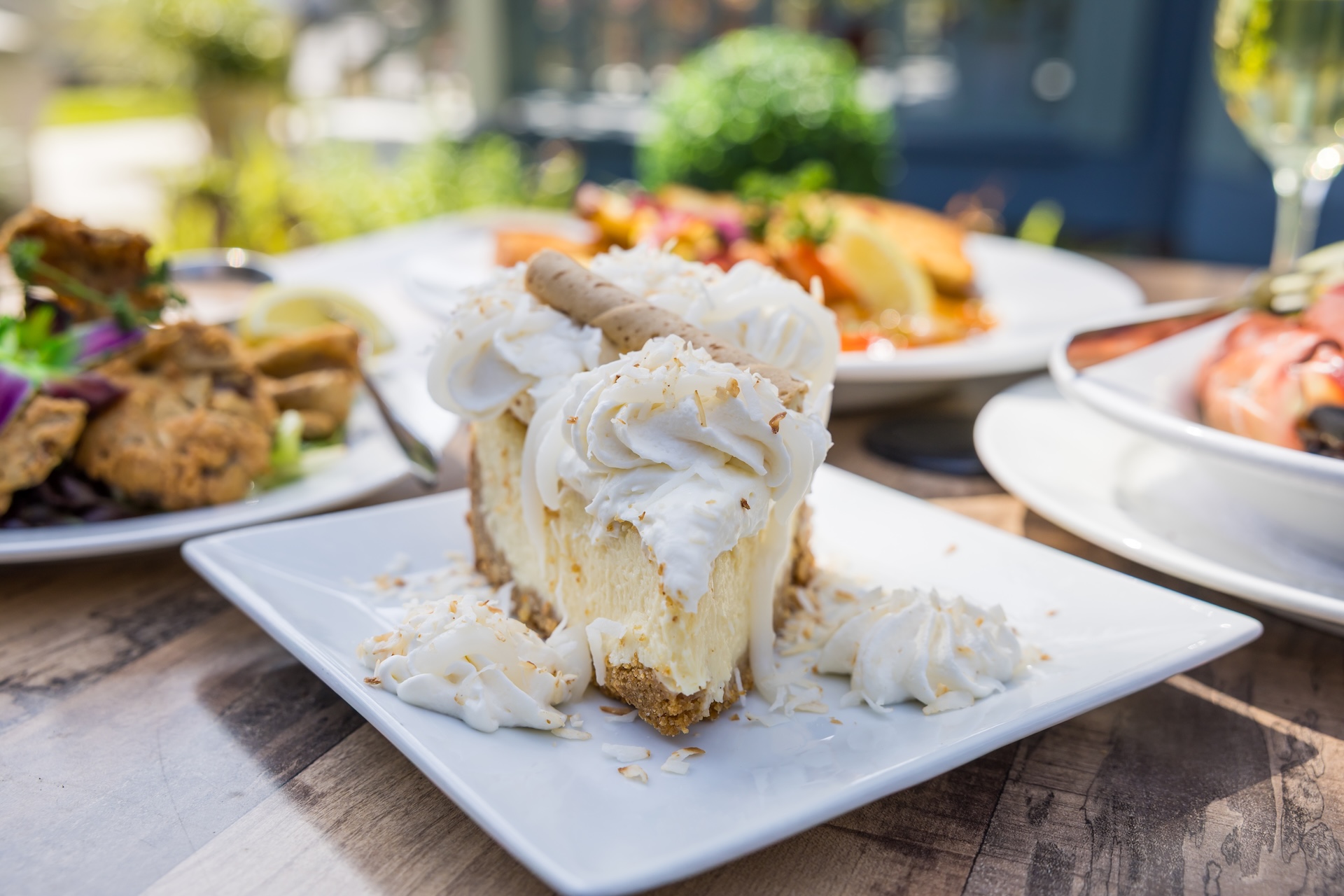 A slice of creamy cheesecake topped with whipped cream and a wafer stick is served on a white plate, garnished with toasted coconut. Other dishes and drinks are blurred in the background on a wooden table.