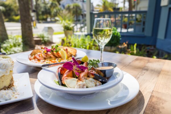 A plate of seafood garnished with flowers and parsley, a glass of white wine, and another dish, all set on an outdoor wooden table with greenery and sunlight in the background.