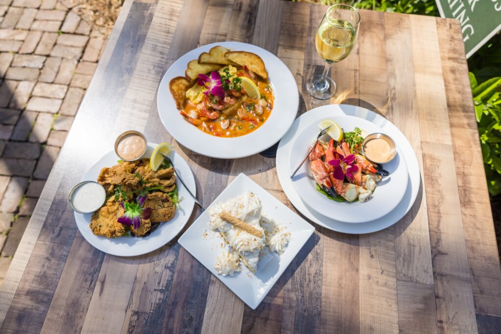 Four plates of gourmet food, including fried chicken, shrimp with toast, seafood with sauce, and a dessert topped with shredded coconut, are arranged on a wooden table with a glass of white wine in sunlight.