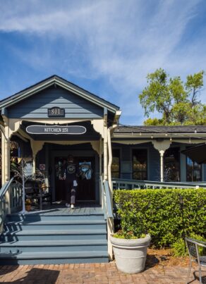 A small blue and gray restaurant with a covered porch, American flag, potted plants, and outdoor seating under a sunny blue sky with wispy clouds.