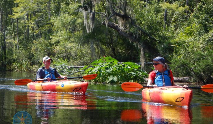 Two people in colorful kayaks paddle on a calm, tree-lined river, wearing life jackets and hats, with green foliage and mossy branches in the background.