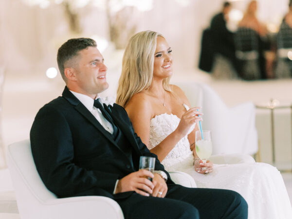A bride and groom dressed in formal wedding attire sit side by side on white chairs, smiling and holding drinks. The background is softly lit with guests seated at tables.