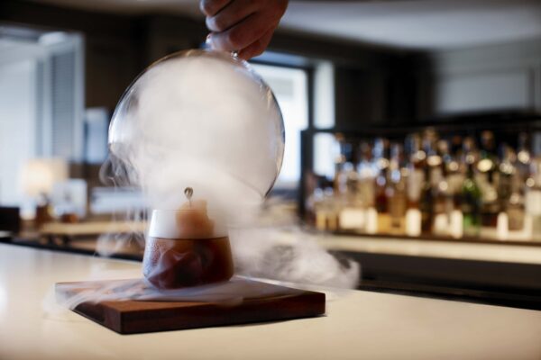 A hand lifts a glass dome releasing smoke from a cocktail on a wooden board at a bar, with bottles blurred in the background.