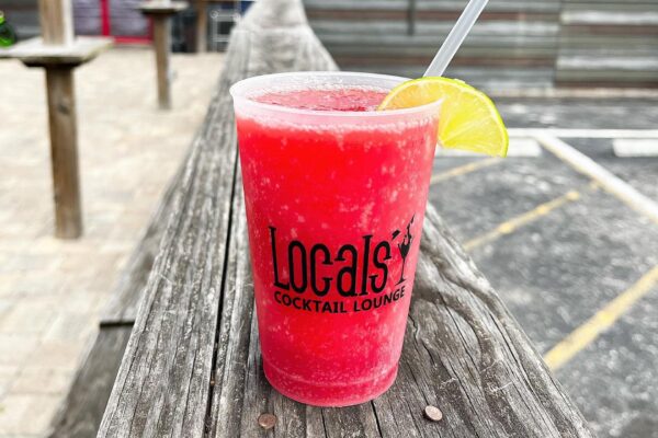 A bright red frozen drink in a clear plastic cup labeled Locals Cocktail Lounge, garnished with a lime wedge and a straw, sits on a wooden railing with a parking lot and rustic outdoor seating in the background.