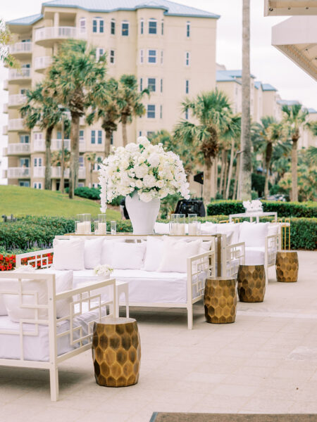 Elegant outdoor lounge area with white sofas and cushions, a large floral arrangement in a white vase, and gold textured side tables, set against palm trees and a beige multi-story building.