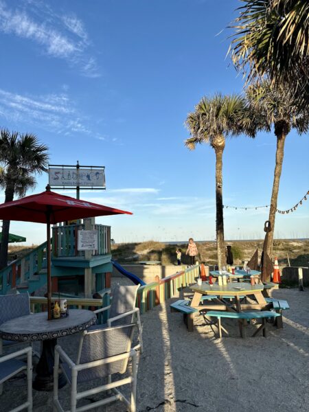 Colorful outdoor seating area at a beachside cafe with picnic tables, palm trees, and a sign reading “SLIDERS.” A person stands on the sandy path near the grassy dunes under a clear blue sky.