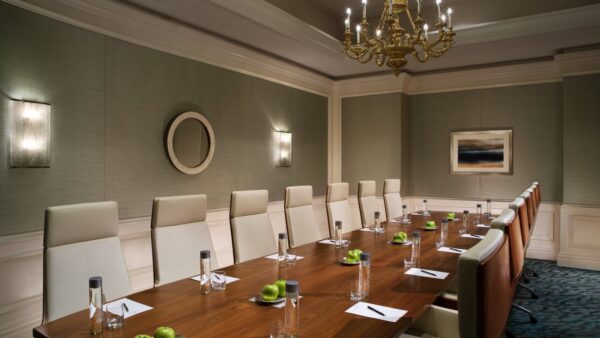 A modern conference room with a long wooden table, white high-back chairs, bottled water, notepads, pens, and green apples, elegant wall decor, and a chandelier overhead.