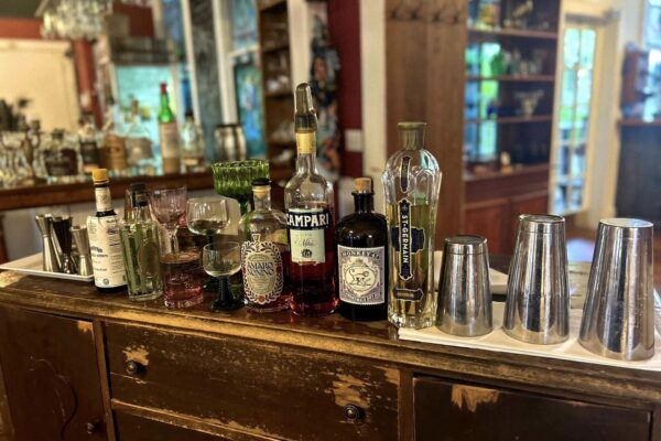 A wooden bar counter displays various bottles of liquor, glassware, and cocktail shakers, with shelves of bottles and glassware in the background. The scene is brightly lit and organized for drink preparation.