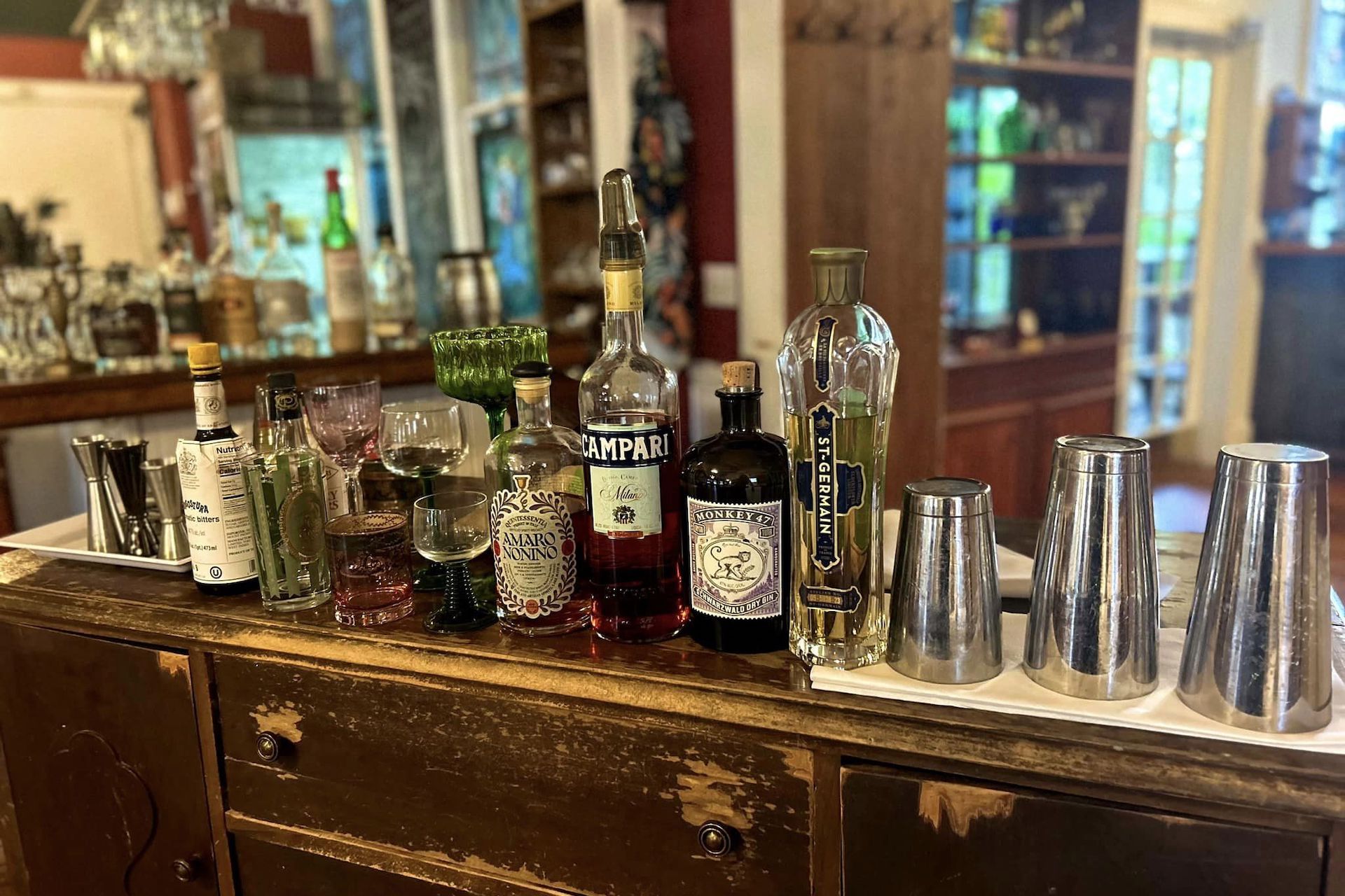 A wooden bar counter displays various bottles of liquor, glassware, and cocktail shakers, with shelves of bottles and glassware in the background. The scene is brightly lit and organized for drink preparation.