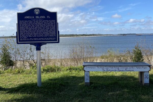A blue historical marker for Amelia Island, FL stands beside a waterfront, with a white bench nearby and a cloudy sky overhead. The water and distant shore are visible in the background.