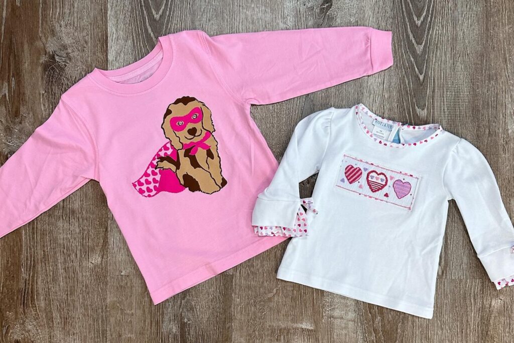 Two long-sleeve childrens shirts on a wooden floor: one pink with a dog in a superhero mask and cape, and one white with a patch of four patterned hearts in pink and red tones.