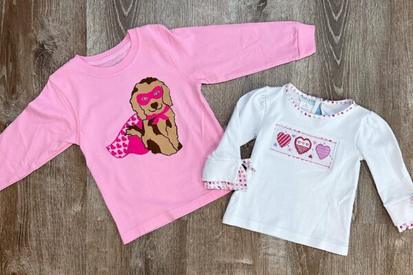 Two long-sleeve childrens shirts on a wooden floor: one pink with a dog in a superhero mask and cape, and one white with a patch of four patterned hearts in pink and red tones.