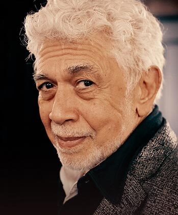 An older man with curly white hair, a trimmed white beard, and mustache, wearing a gray blazer over a black shirt, looks over his shoulder and smiles softly against a dark background.