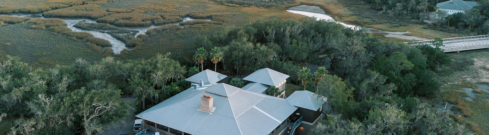 Aerial view of a house with a swimming pool surrounded by trees and marshland; nearby houses and winding waterways are visible in the background under a sunset sky.