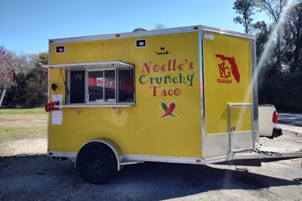A yellow food truck with Noelles Crunchy Taco written on the side, two chili peppers graphic, and an OPEN sign in the window. The truck has a Florida state outline and is parked outdoors on a sunny day.