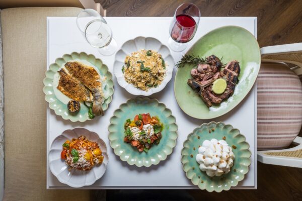 A white table set with six plated gourmet dishes, including fish, pasta, steak, salad, and a meringue dessert, along with a glass of red wine and an empty wine glass.