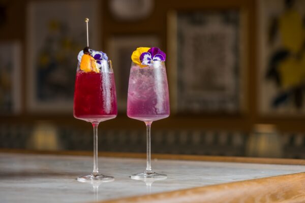 Two colorful cocktails in stemmed glasses sit on a marble countertop. One is deep red with fruit garnish; the other is light purple with ice and edible flowers. The background is softly blurred with framed art on the walls.