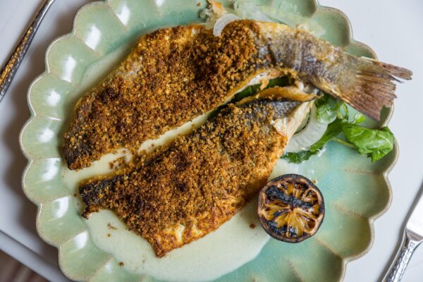 Two pieces of breaded and baked fish fillets on a green scalloped plate, served with a charred lemon half and some leafy greens on the side.