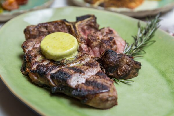 A grilled steak topped with a round pat of herb butter, garnished with a sprig of rosemary, served on a light green plate.