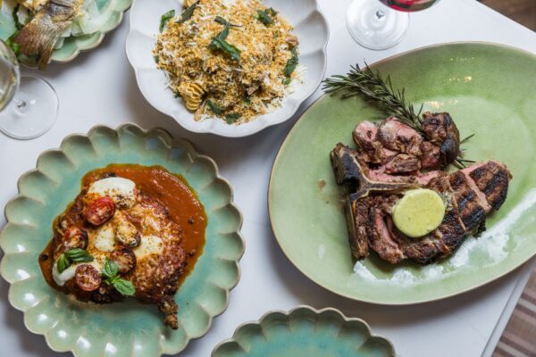 A table set with Italian dishes, including grilled steak with herb butter, pasta with cheese and basil, and eggplant parmesan with tomato sauce, garnished with fresh herbs, alongside glasses of white and red wine.