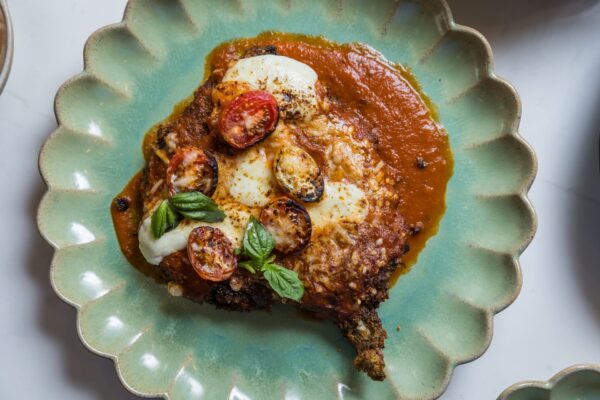 A breaded, baked meat cutlet topped with melted cheese, sliced cherry tomatoes, fresh basil, and tomato sauce, served on a scalloped, pale green plate.