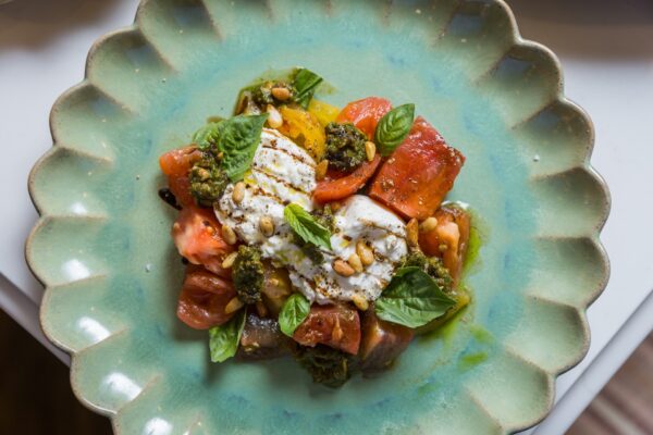 A colorful salad on a green scalloped plate, featuring sliced tomatoes, fresh basil leaves, creamy burrata cheese, pine nuts, and a drizzle of green pesto.