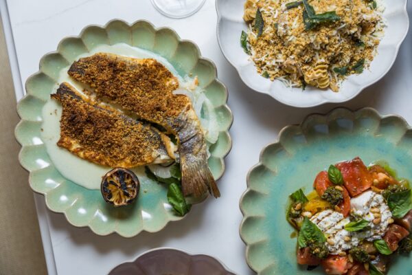 A table with three plates: breaded fish with sauce and grilled lemon, pasta with breadcrumbs and herbs, and a salad with tomatoes, chickpeas, basil, and cheese.