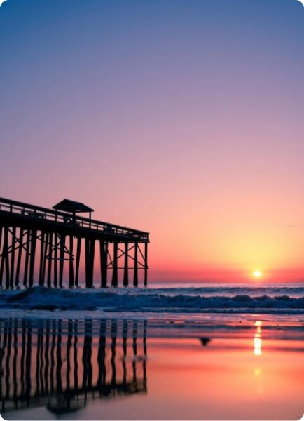 A wooden pier extends over the ocean at sunset, with vibrant pink and purple skies reflected in the wet sand and gentle waves rolling onto the shore.