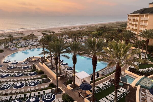 A resort by the beach at sunset, featuring palm trees, two large outdoor pools, hot tub, numerous lounge chairs, and a sandy shore with gentle waves in the background.