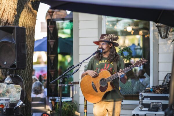 A man with long hair and a feathered hat plays an acoustic guitar and sings into a microphone at an outdoor event, standing near a building with glass windows and surrounded by music equipment.