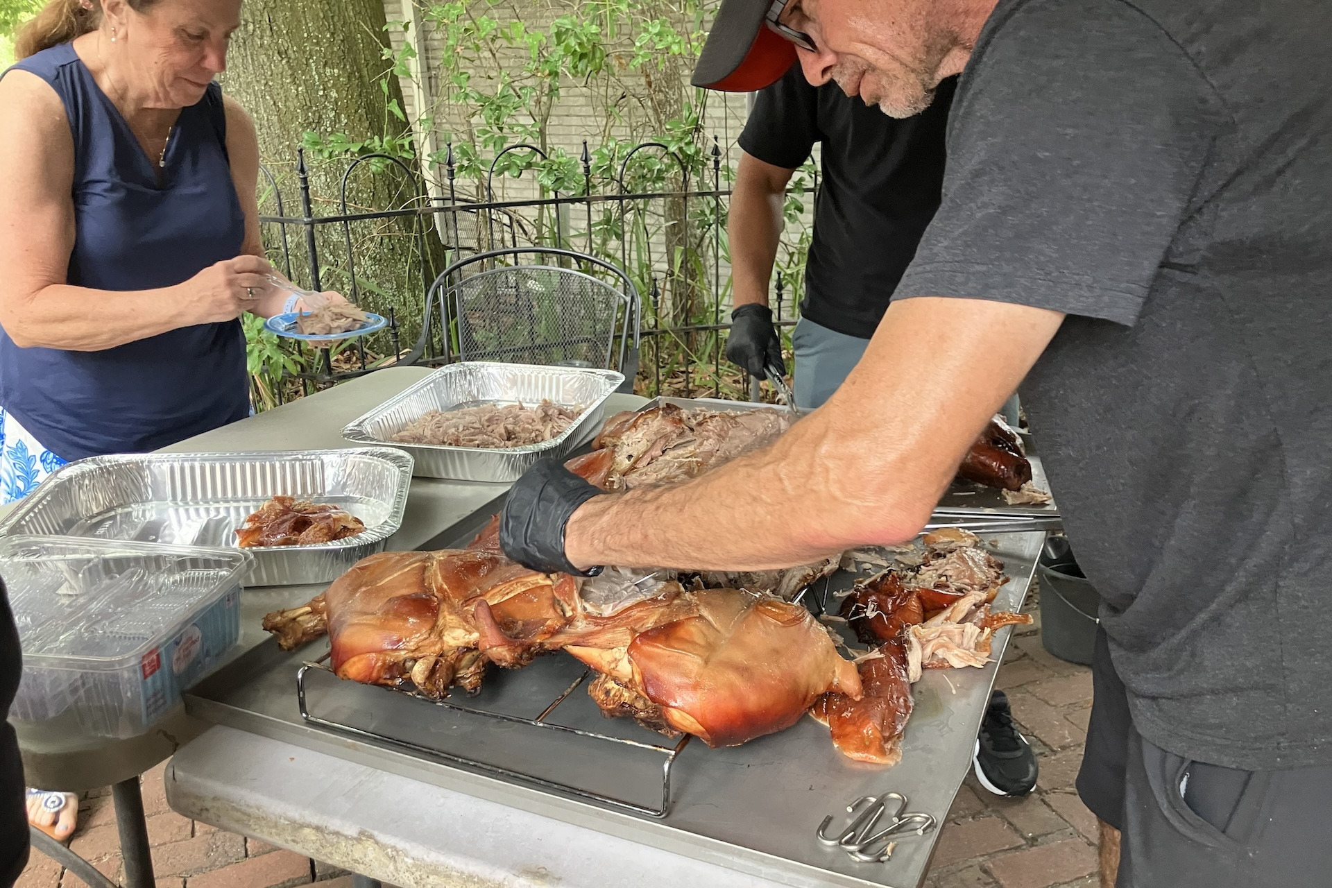 Three people are shredding and serving a roasted pig on a table outdoors. Metal trays are being filled with pulled pork. The setting appears casual, with trees and a fence in the background.