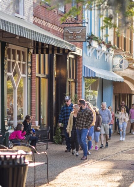 People walk along a sunny, tree-lined sidewalk with storefronts and awnings. Some sit at outdoor tables, while others stroll in groups, enjoying the lively street scene.