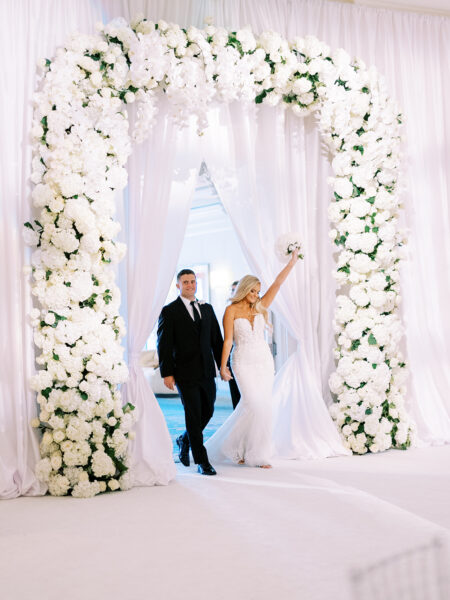 A bride in a white dress and a groom in a black suit walk under a large, white floral archway. The bride raises her bouquet triumphantly while they both smile, surrounded by draped white fabric.