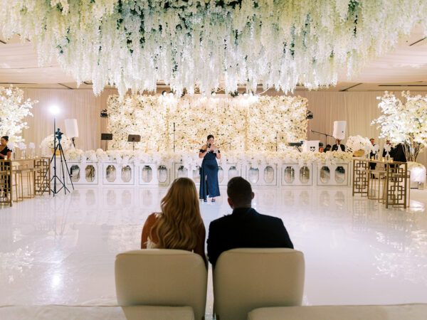 A bride and groom sit together facing a glitzy wedding reception ballroom, decorated with hanging white flowers and elegant white floral arrangements, as a woman gives a speech at the front.