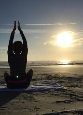 A person sits cross-legged on a beach towel at sunrise, arms raised overhead, silhouetted against the sun and ocean waves.