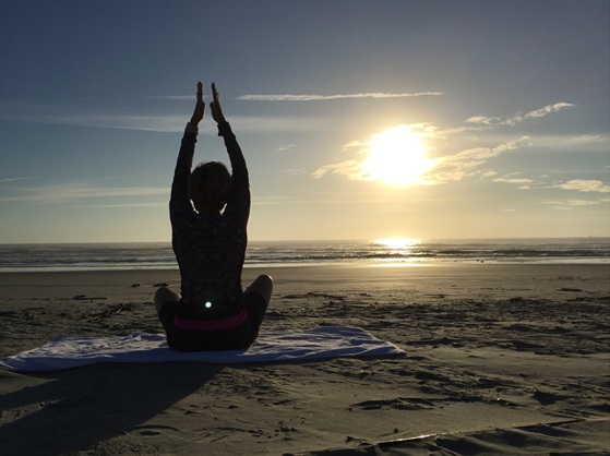 A person sits cross-legged on a beach towel at sunrise, arms raised overhead, silhouetted against the sun and ocean waves.