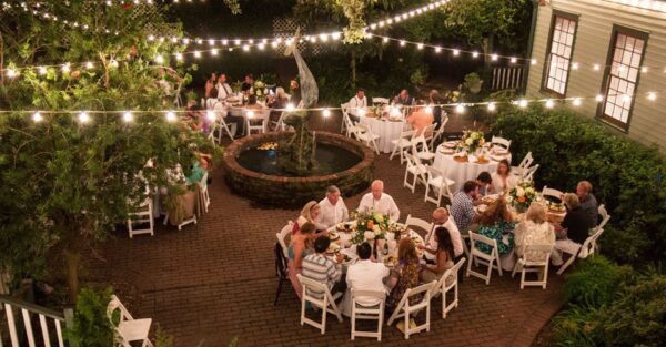 A nighttime outdoor gathering with people sitting at round tables covered in white cloths, surrounded by greenery and string lights, with a fountain in the center. The atmosphere is festive and intimate.