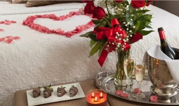 A romantic setup with a bed decorated with a heart of rose petals, a bouquet of red roses, two champagne glasses, chocolate-covered strawberries, a lit candle, and a bottle of wine in an ice bucket.