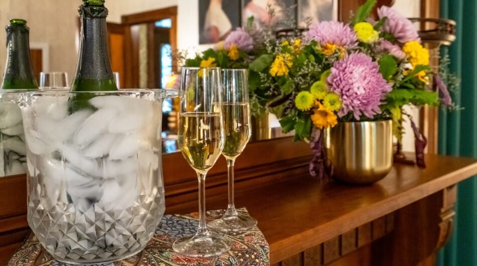 A bottle of champagne in an ice bucket and two filled champagne glasses sit on a patterned cloth atop a wooden mantel, next to a bouquet of colorful flowers in a gold vase.