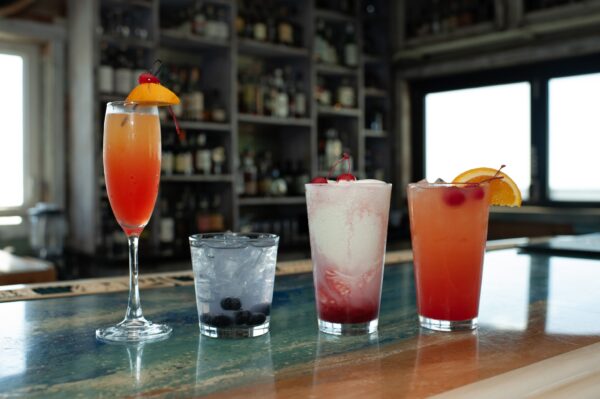 Four colorful cocktails sit on a bar counter, each garnished with fruit such as cherries, blueberries, and orange slices, with bottles of liquor visible on shelves in the blurred background.