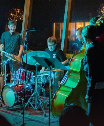 A jazz band performs on stage with a drummer, upright bass player, and other musicians. The scene is dimly lit, creating a cozy, intimate atmosphere. The musicians appear engaged and focused on their music.
